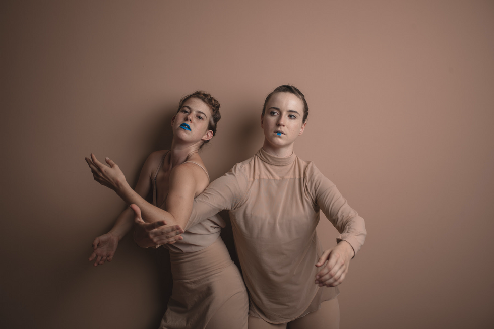 Two dancers wearing bright blue lipstick and beige clothing that matches the backdrop pose together, contracting towards their centers. One's elbow awkwardly hooks the other's, their hips knocking together as they gaze mistrustfully around.