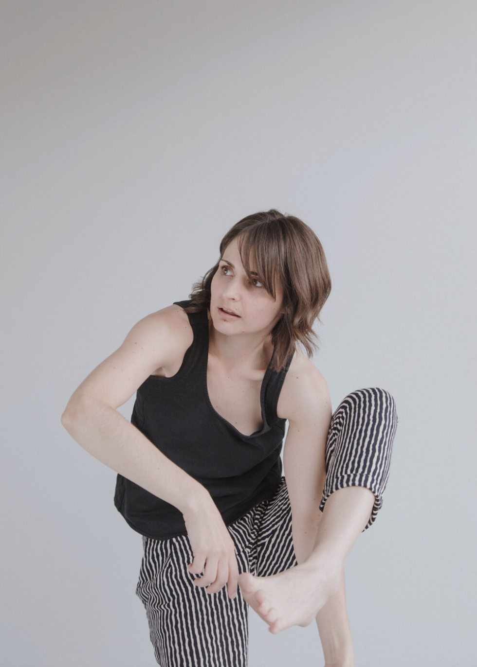 Elisabeth Motley, a white woman with brown eyes, chin-length layered brown hair with bangs, dances in front of a light gray backdrop. She wears a sleeveless black shirt and black and white thin-striped ankle-length pants and is barefoot. She stands on her right leg with her left leg hooked in a front attitude, and she crouches asymmetrically so as to touch the side of her right index finger to the base of her left big toe. Her left arm reaches down through the hole created by her right arm and left leg. She looks up on a shallow diagonal to the right, lips slightly parted.