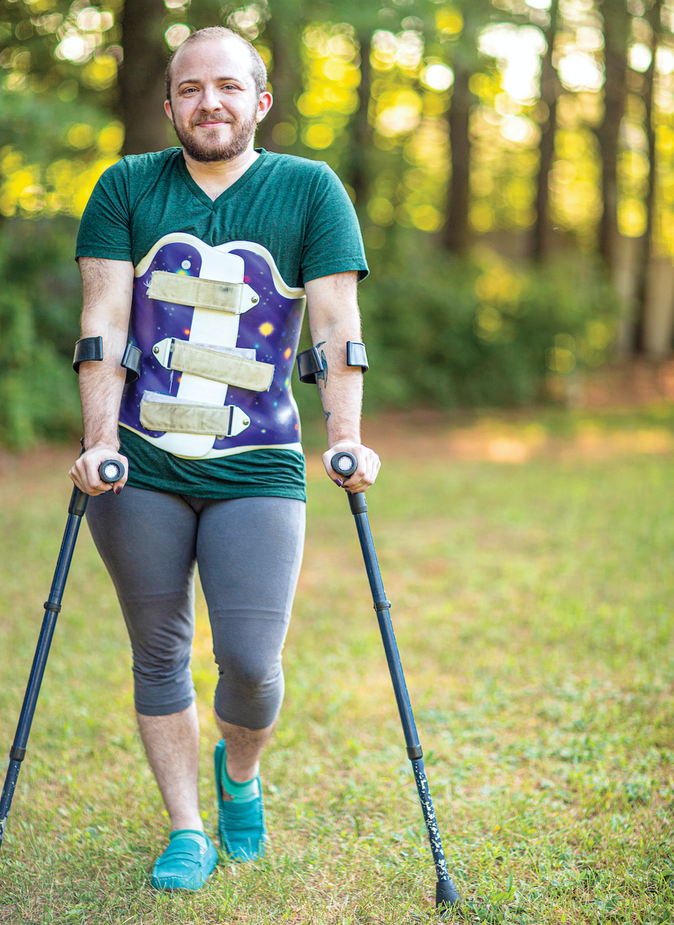 Toby MacNutt stands with crutches and a back brace, on a green landscape