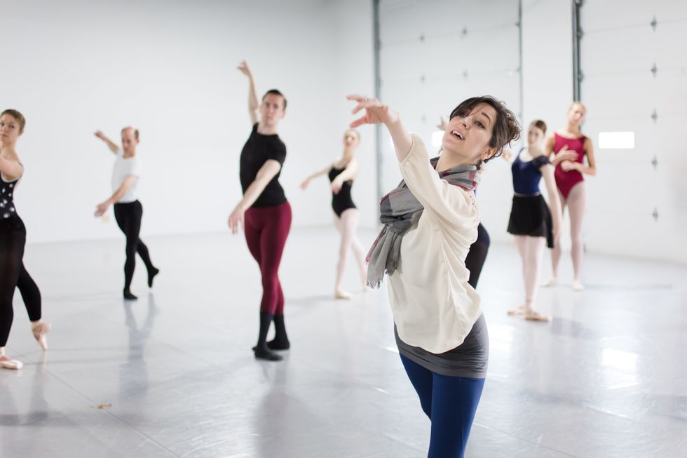 Slager teaches a ballet class to a group of adults, demonstrating with her arm outstretched in the front of the light-filled classroom