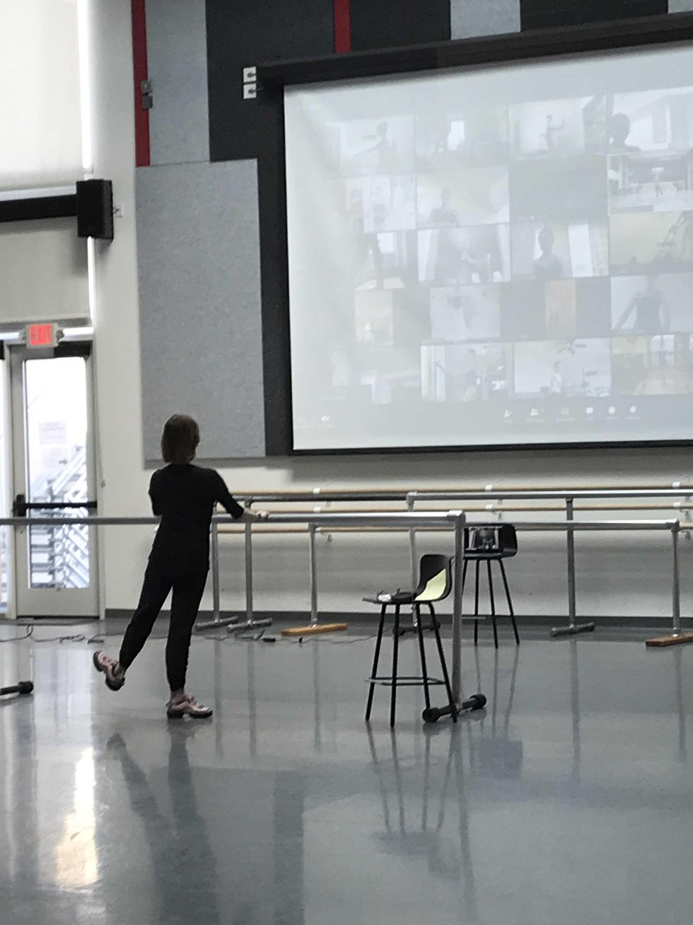 A woman alone in a studio at barre demonstrates a tendu flex looking up at a projection of students on Zoom.