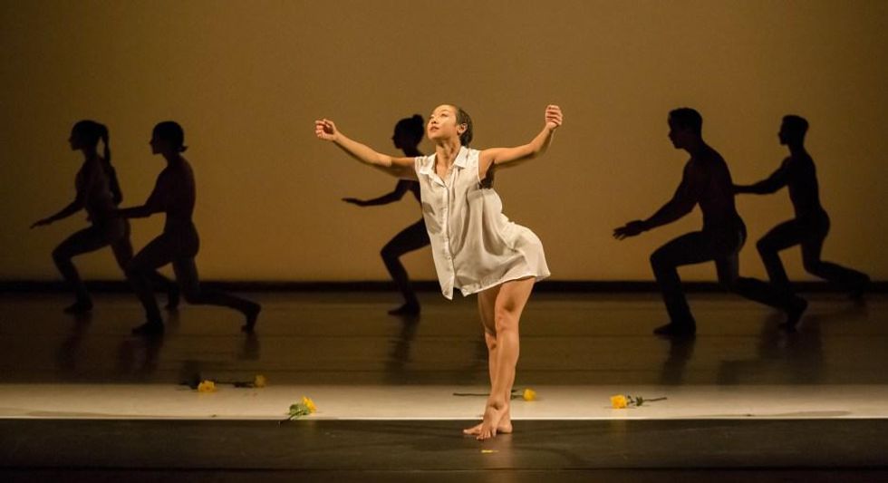 Ching Ching Wong holds her arms open to the audience, one leg pointed, on a stage with dancers silhouetted behind her, yellow roses scattered on the floor