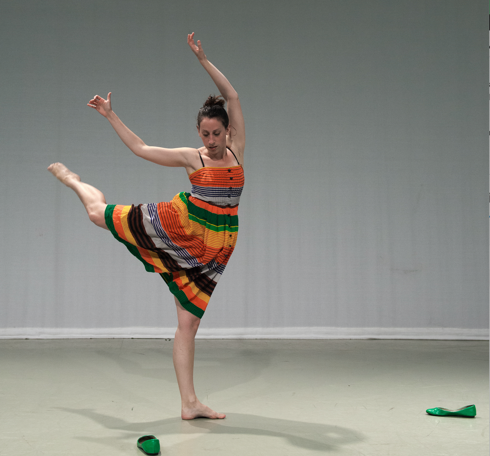 Monica Thomas in a multi-colored dress stands barefoot in attitude back, two green shoes scattered on the floor near her.