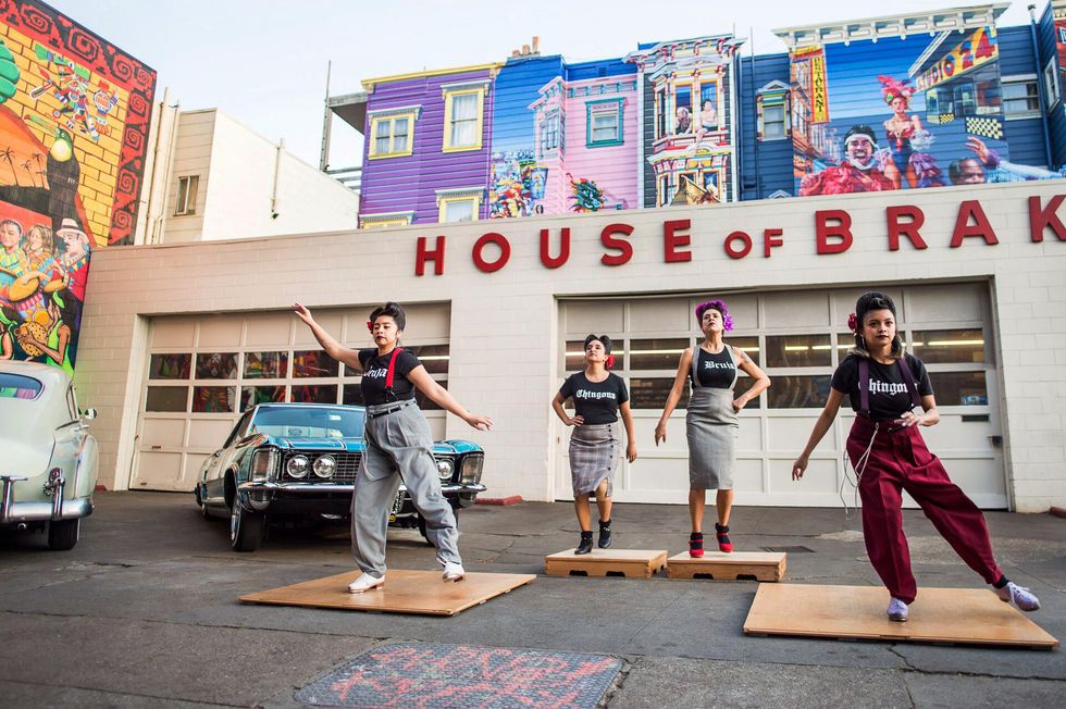 Four women tap on wooden planks in front of a garage with the words "House of Brakes"
