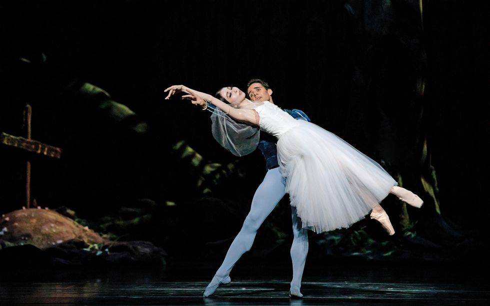 Yuriko Kajiya lays almost horizontally across Connor Walsh's arms, in a long white romantic tutu and pointe shoes