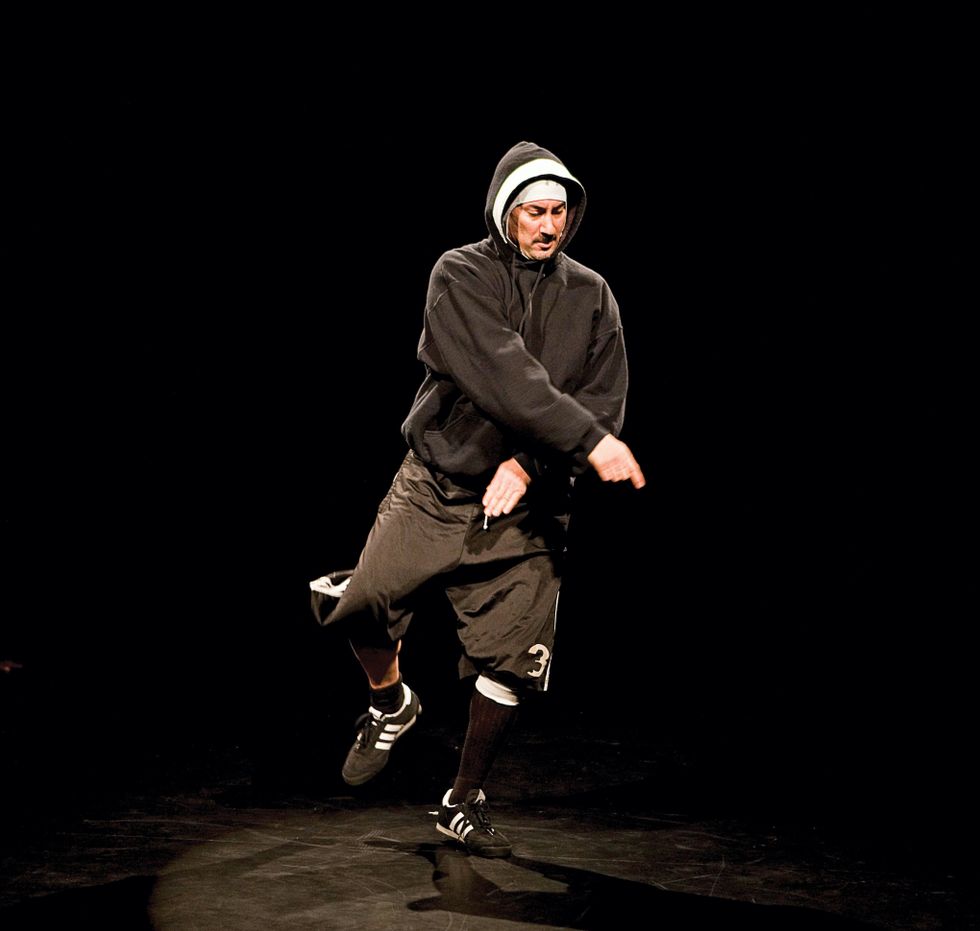 Doug Elkins in a black hoodie over his head, athletic pants and sneakers, crosses his arms, looking down on a black stage