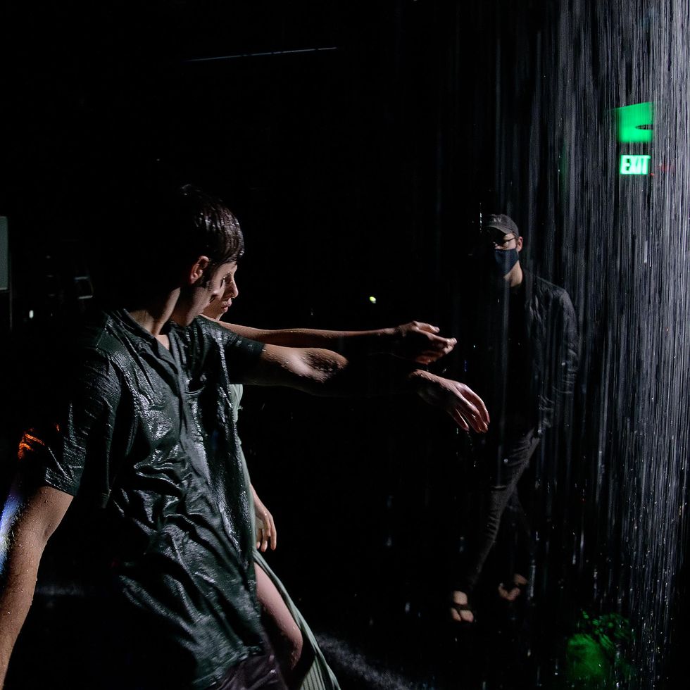 A stream of water falls down on two dancers while Bell, in a mask, watches from the background