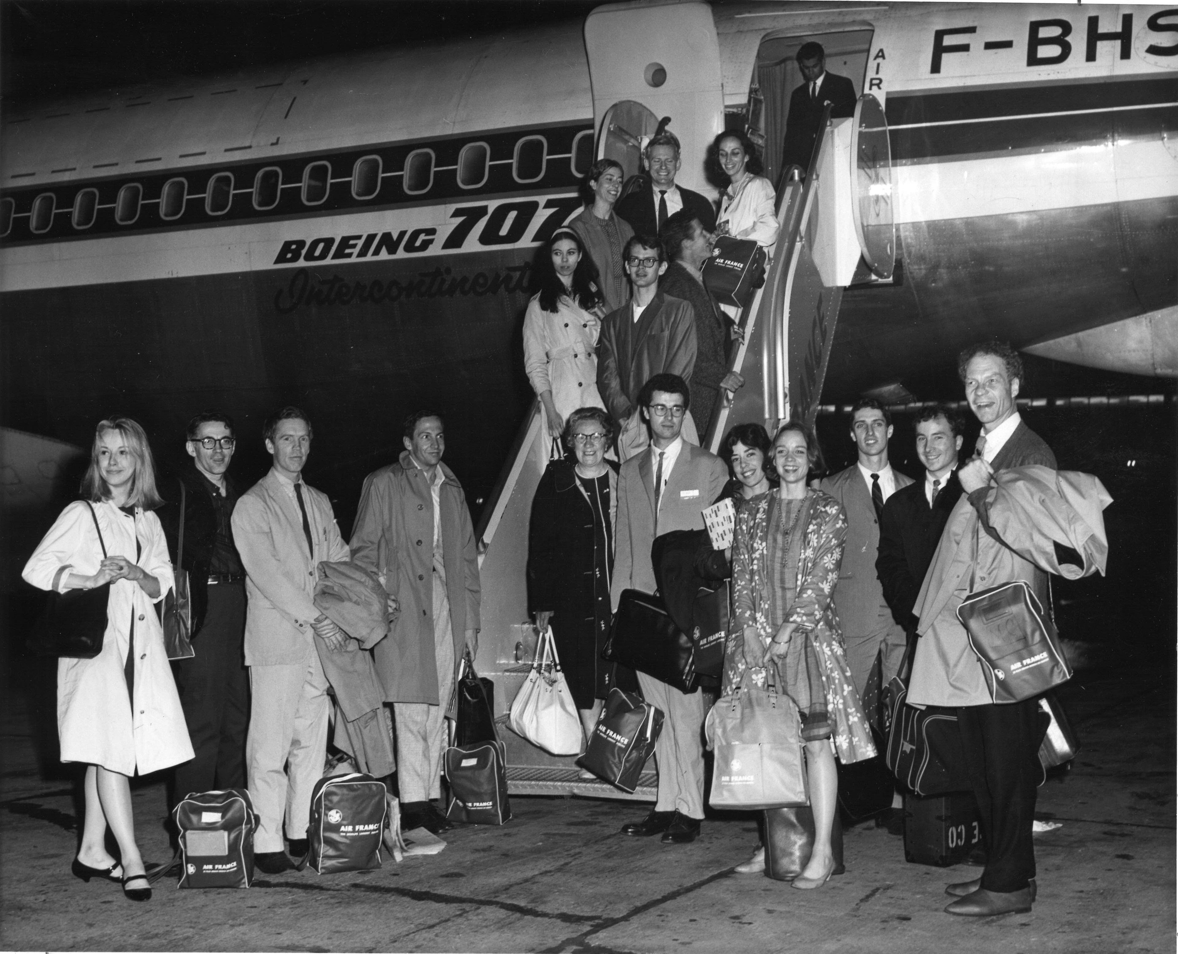 A grinning Merce Cunningham, holding a coat and travel bag, stands with his company by the boarding steps to a Boeing 707. The women wear dresses, the men ties.