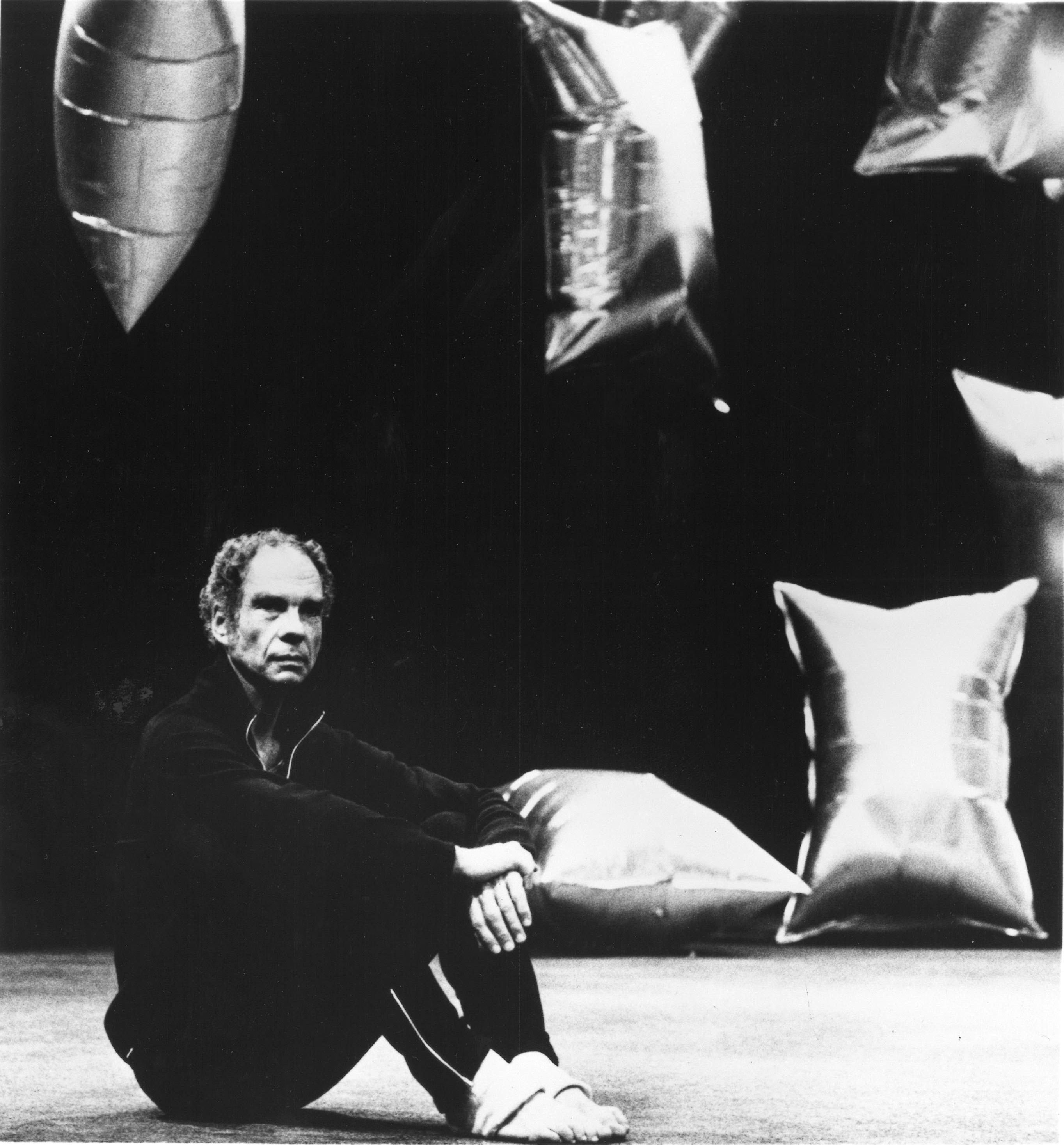 Merce Cunningham, in a black tracksuit, sits with his knees pulled up onstage. Behind him, the silver balloons designed by Andy Warhol for Cunningham's RainForest.