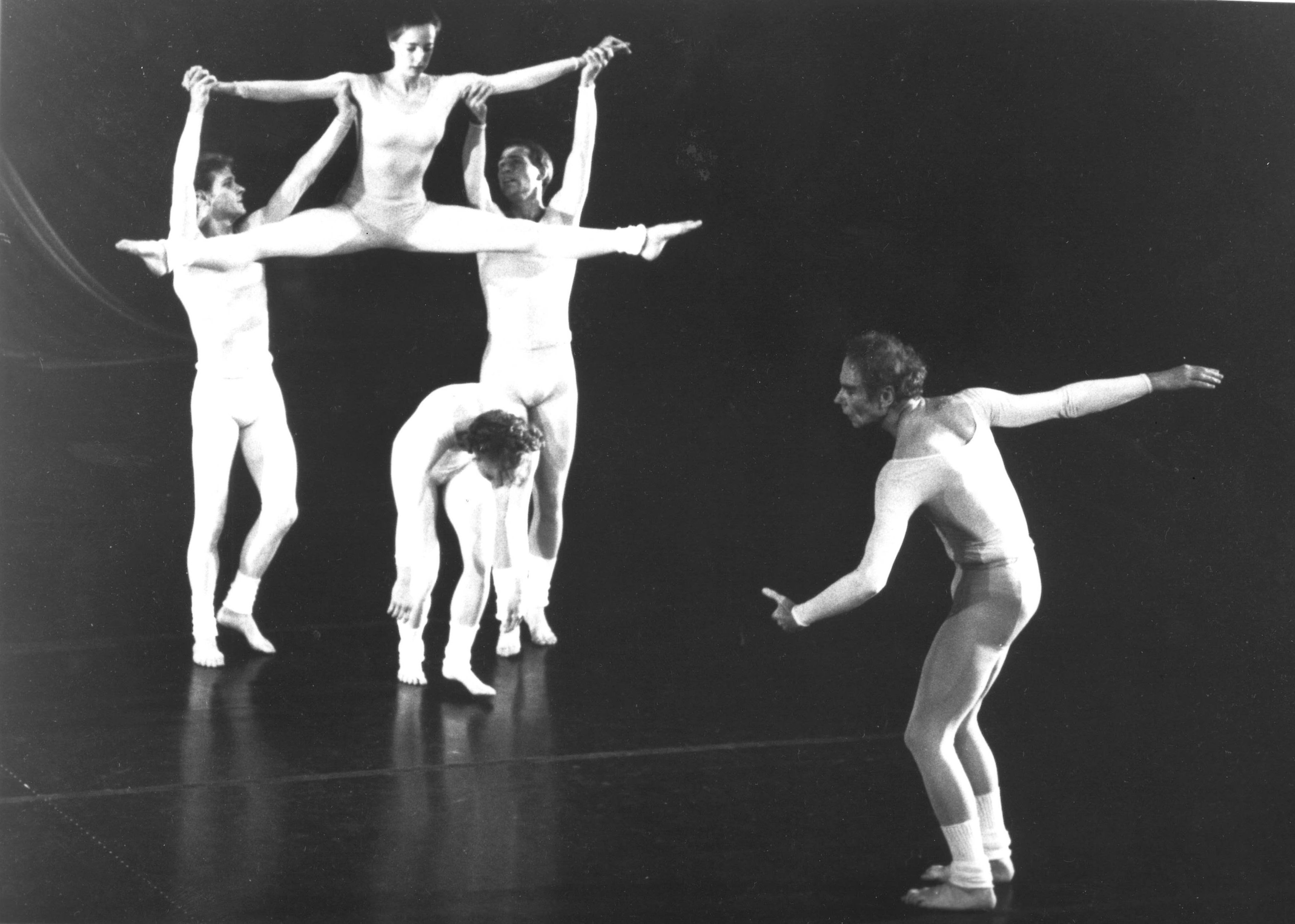 Two male dancers list a woman, legs split, over a third, who is crouching. Merce Cunningham gestures with an arm behind him in the corner. All wear pale unitards and legwarmers.
