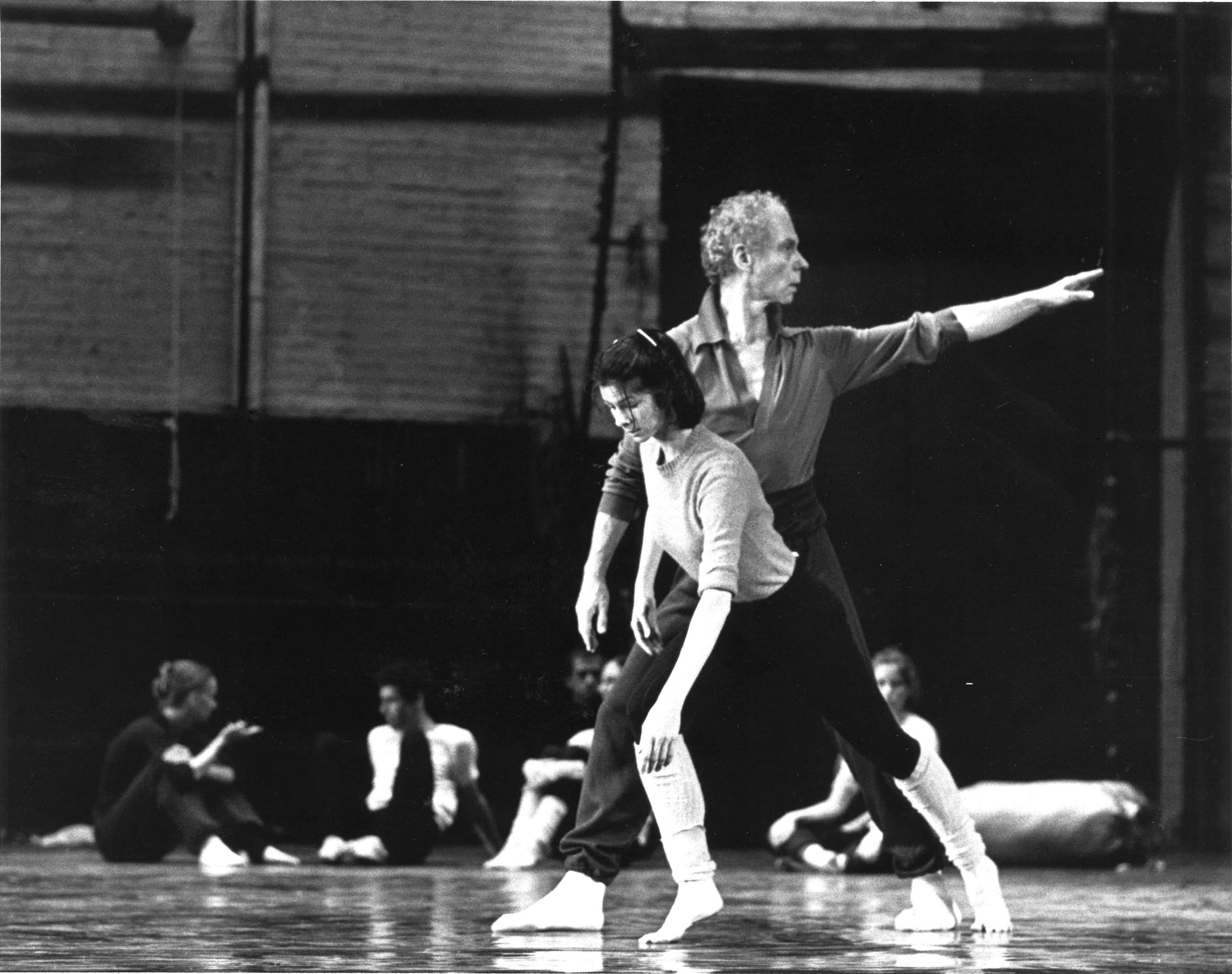 Catherine Kerr gazes down as she balances in a forced arch lunge; Merce Cunningham, just behind her, mirrors her lunge while looking over his back shoulder. Out of focus in the background are seated dancers, chatting and watching. All wear rehearsal clothes.