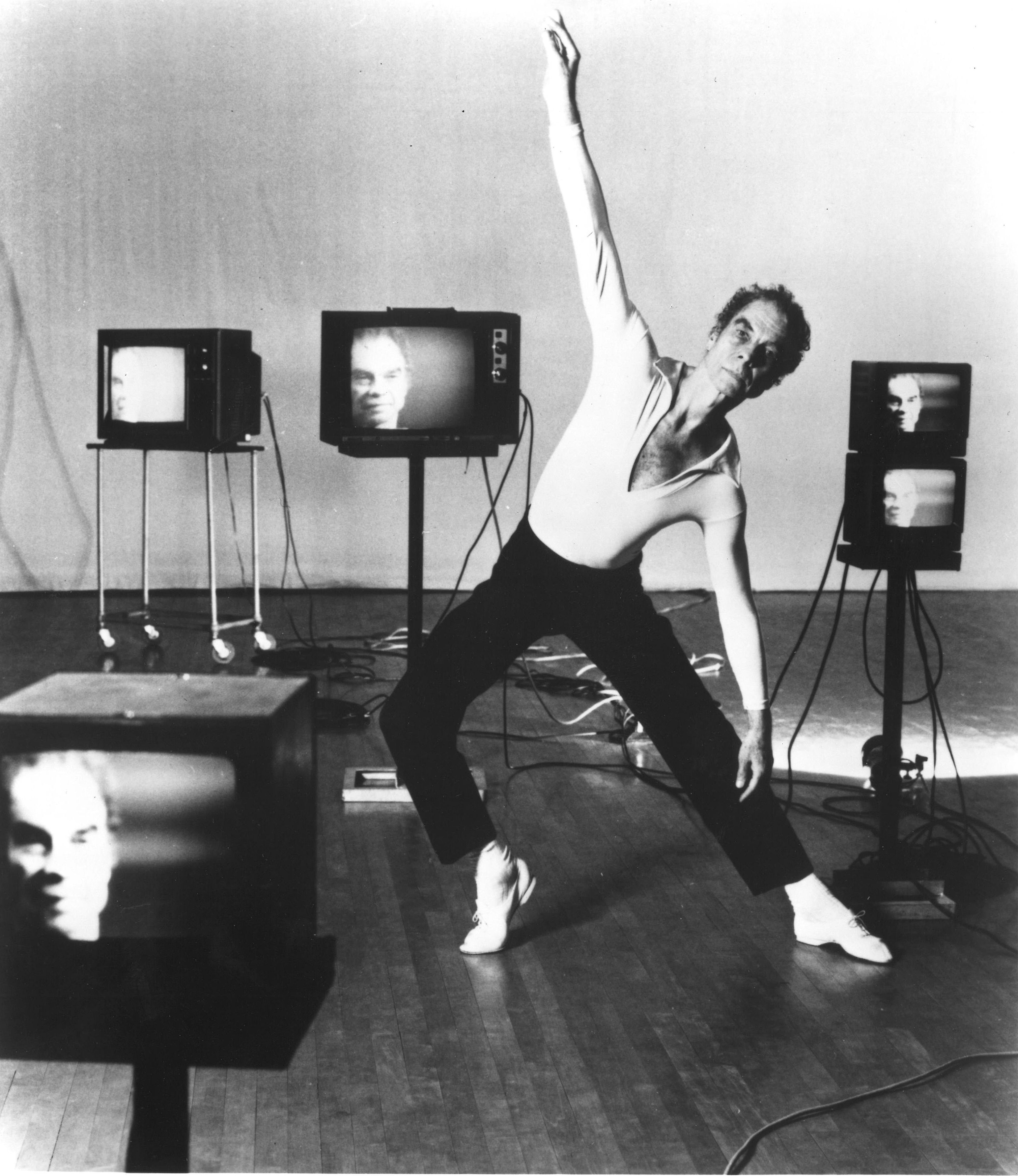 Five small, square television sets of various sizes show a blurry image of Merce Cunningham's face. Cunningham himself is in the middle of the seemingly random array, holding a side lunge with his right arm raised and right foot in forced arch.