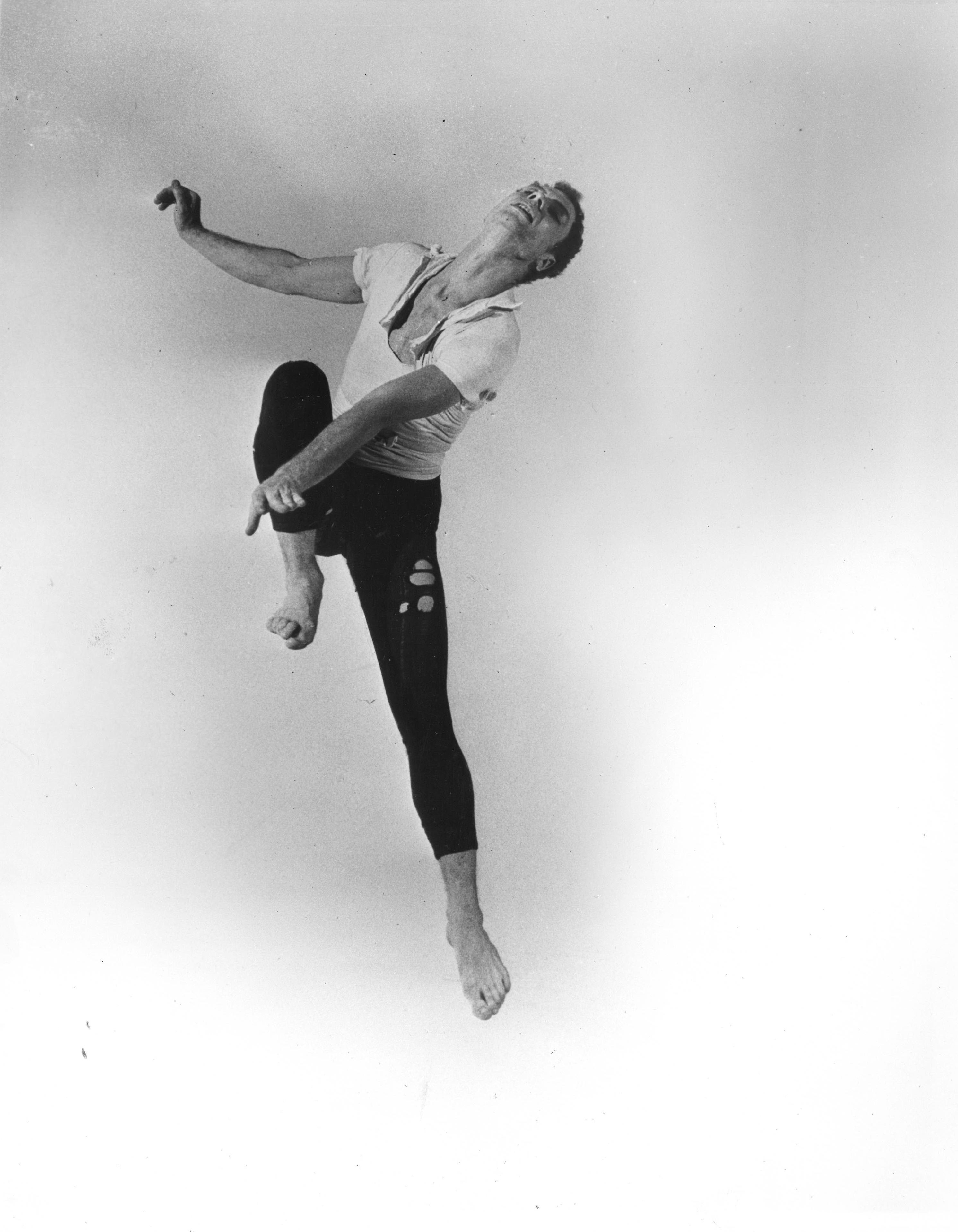 A young Merce Cunningham, dressed in hole-filled rehearsal-wear, jumps, one knee raised to his chest, head flung back.