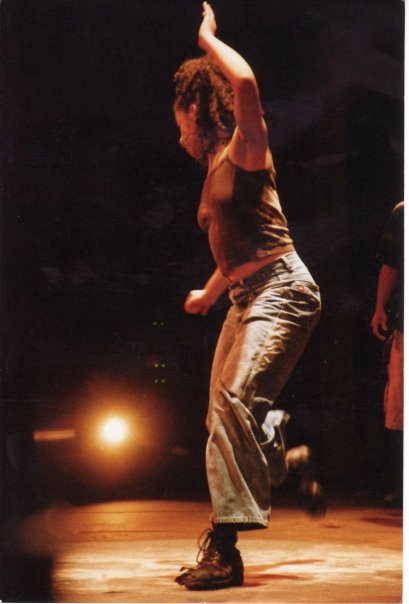 A shot from the wings shows Kamal Sinclair in a blur of motion onstage, her face away from the camera as her curls fly. Her left hand flies up by her head as she stomps onto that foot; her right foot a blur kicked up by her ankle.