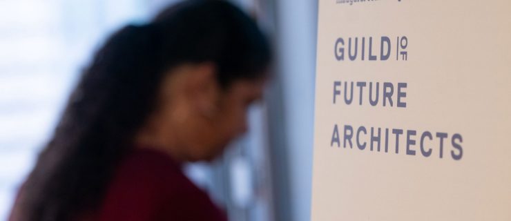 A sign reads "Welcome to our inaugural convening: Guild of Future Architects." Blurry in the background, a woman with curly dark hair is scene in profile as she moves through the door.