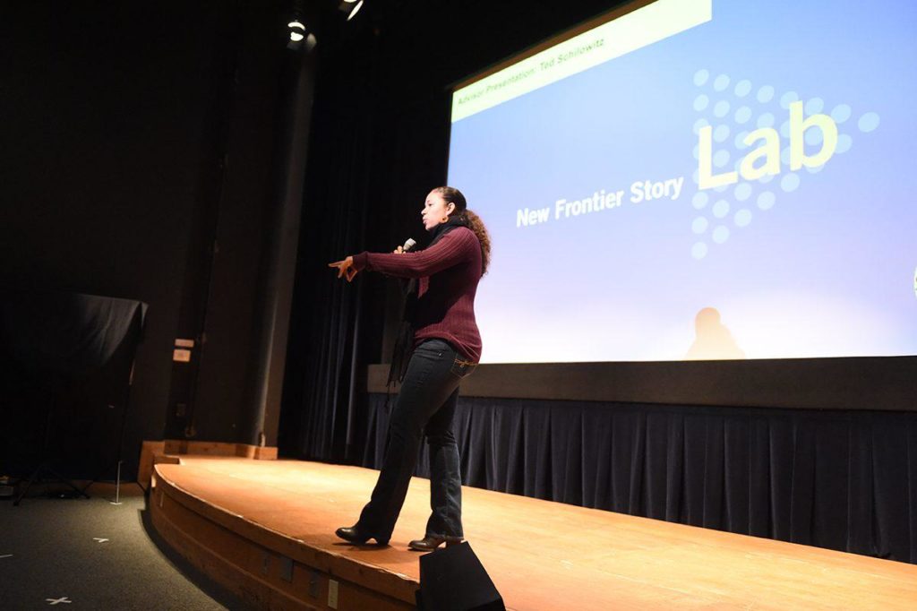 Kamal Sinclair, in jeans and a sweater, holds a microphone as she speaks on a narrow stage. The screen behind her bears a blue and white logo reading, "New Frontier Story Lab."