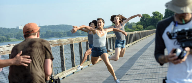 Three dancers on a wooden pier jump towards a camera, blue sky and water in the background