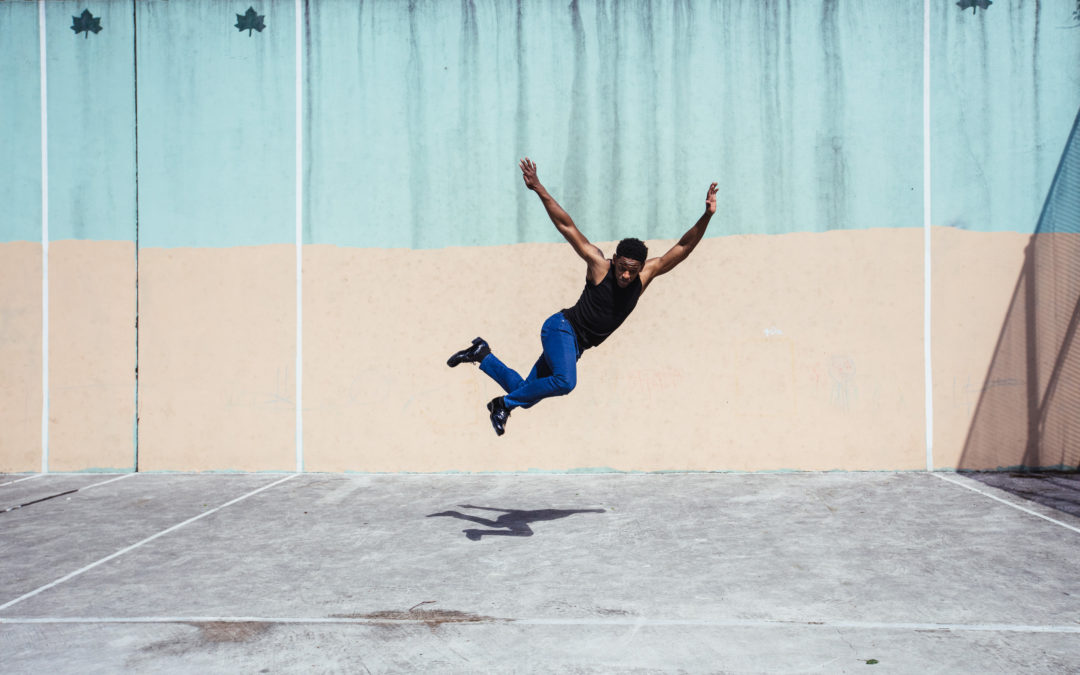 Jared Grimes, wearing black tap shoes, skinny jeans, and a black tank top, flies above a stretch of pavement, looking down at his shadow.