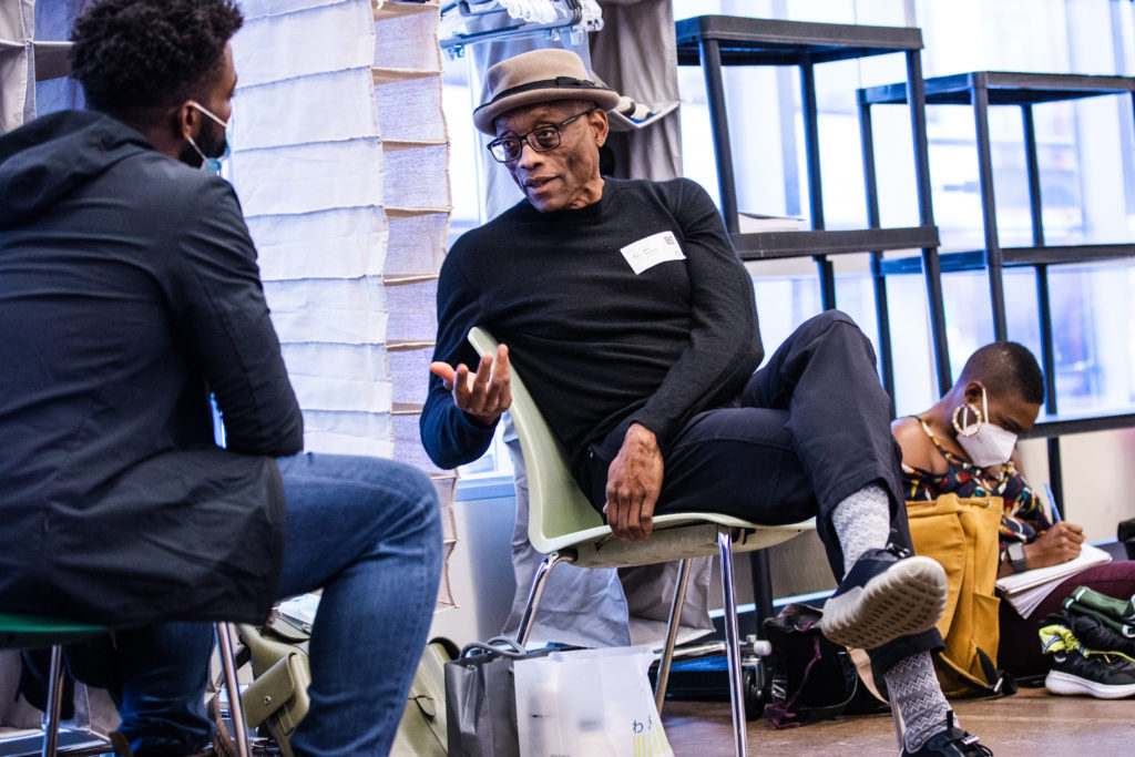 Bill T. Jones leans back in his chair, gesturing with one hand as he turns to speak with a person whose back is to the camera. Various bags and rehearsal detritus line the space. A masked Black woman sits on the floor in the background, writing notes.
