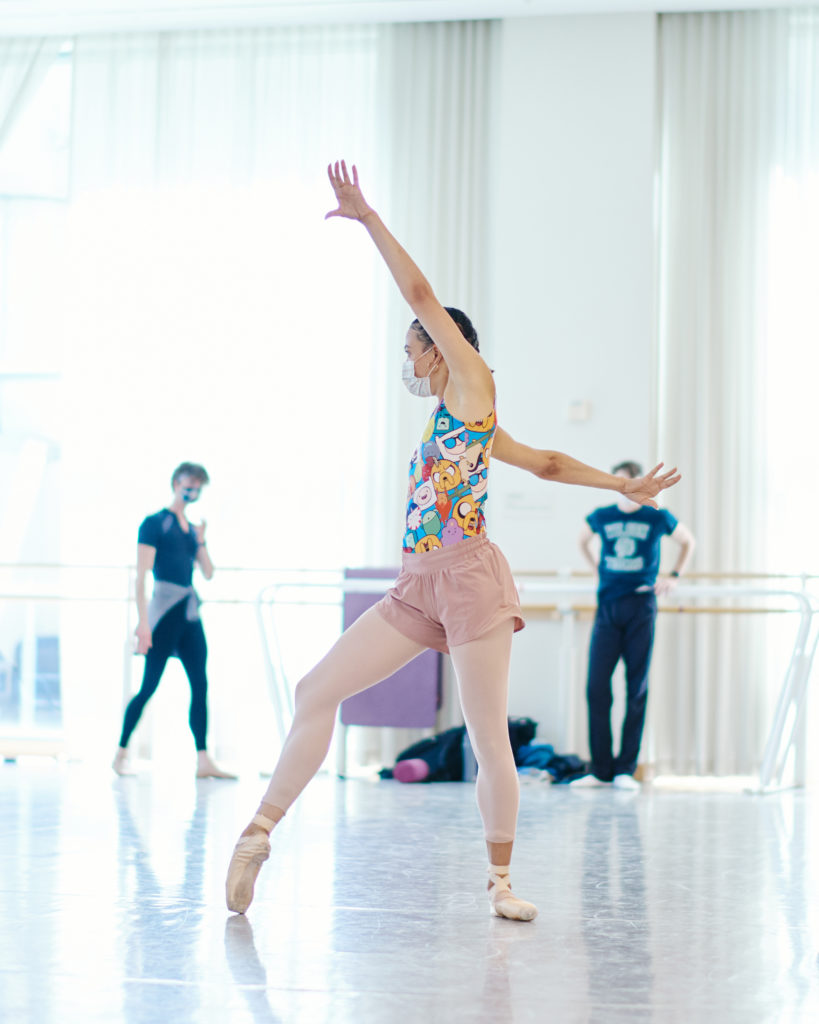 In a sunny studio, Nikisha Fogo, wearing pointe shoes, pink tights and shorts, and a colorful leotard, poses in profile to the camera. Her right shoe is dug into the ground in forced arch, opposite arm flying up by her head while the other splays behind.
