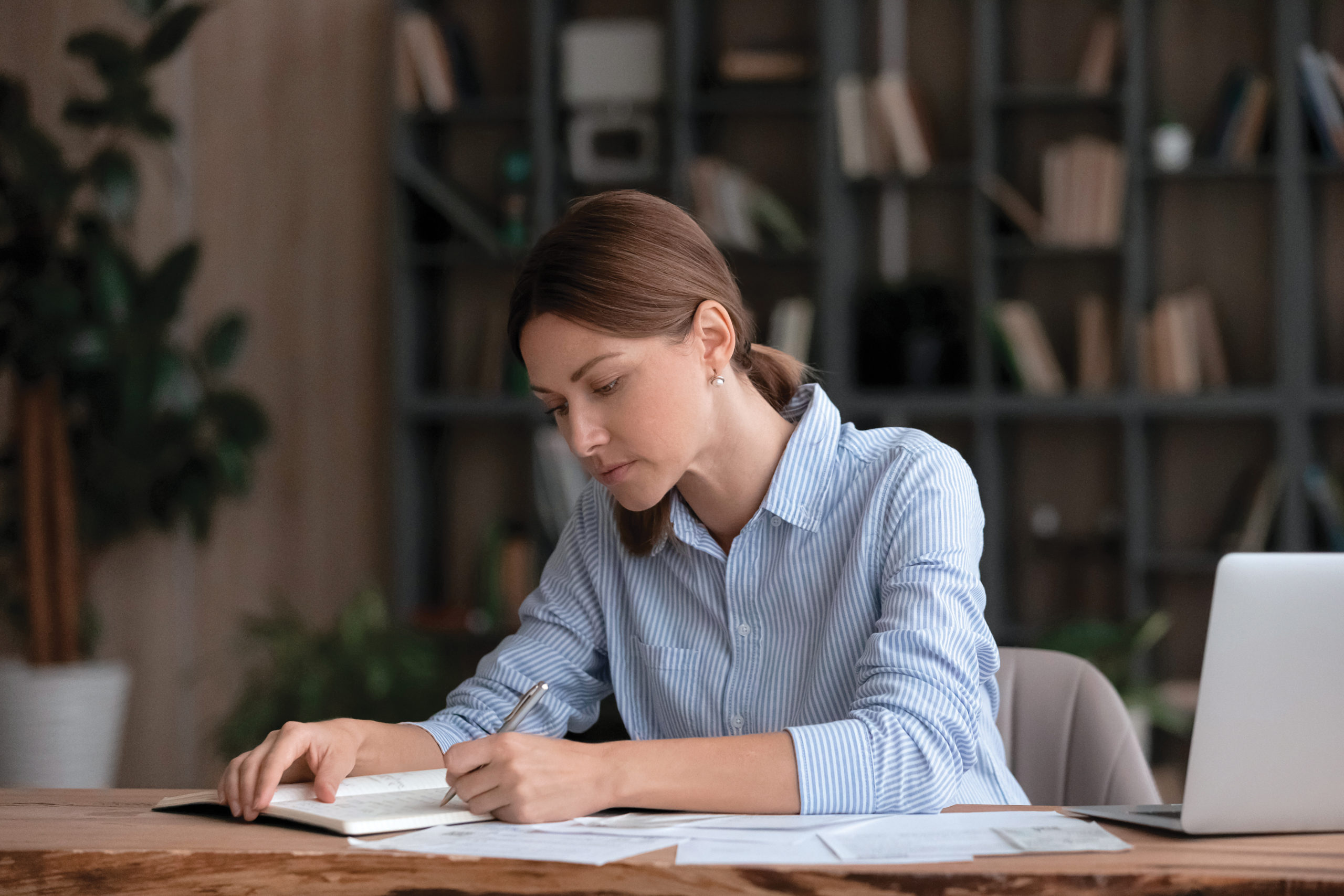 Concentrated young 30s caucasian woman writing business notes in paper organizer, planning future investments or managing monthly budget, reviewing bills or taxes, sitting at table at home office.