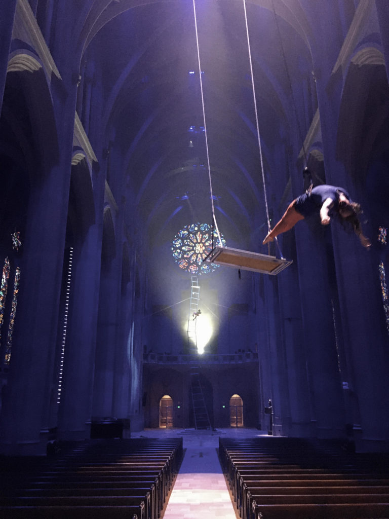 In a soaring, purple-lit cathedral dotted with stained glass windows, an aerialist arches back, parallel to the ground as she flies away from a wooden swing; both are suspended at least 20 feet over the ground. In the background, another aerialist climbs a precarious looking ladder.