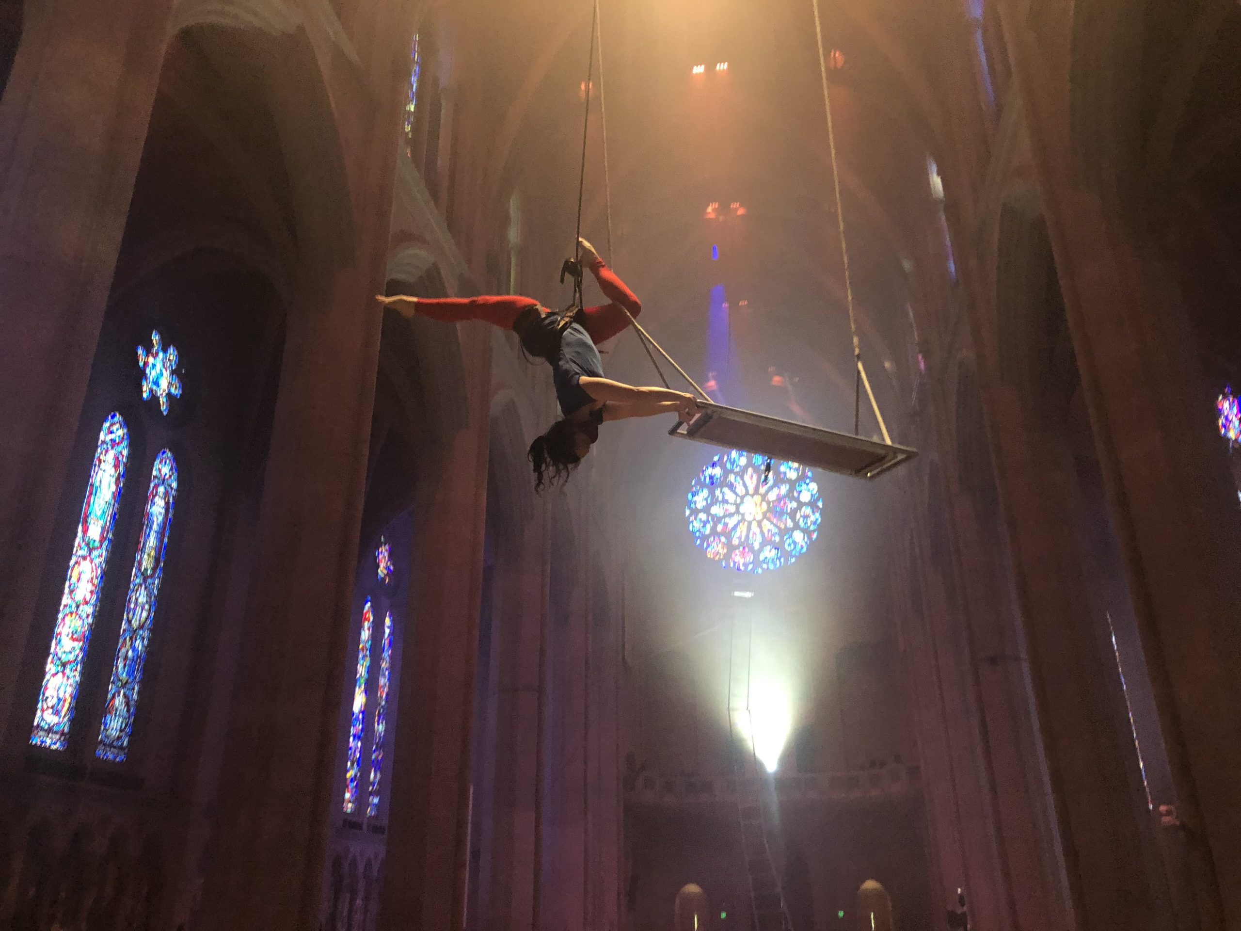 In a soaring cathedral space dotted with stained glass windows, a dancer is suspended, upside down, from the ceiling. She holds a wooden swing with both hands, back slightly arched and knees bent into an upside-down stag leap.