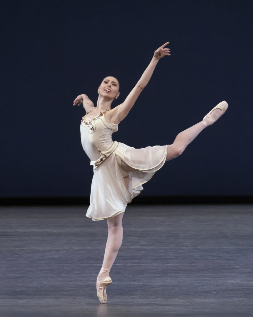 Isabella LaFreniere is alone onstage in a high, open back attitude en pointe, her working arm mirroring the angle of her leg, the other extending long to the side. She wears pink tights and pointe shoes with an off-white, knee-length dress, the skirt slit to the waist.