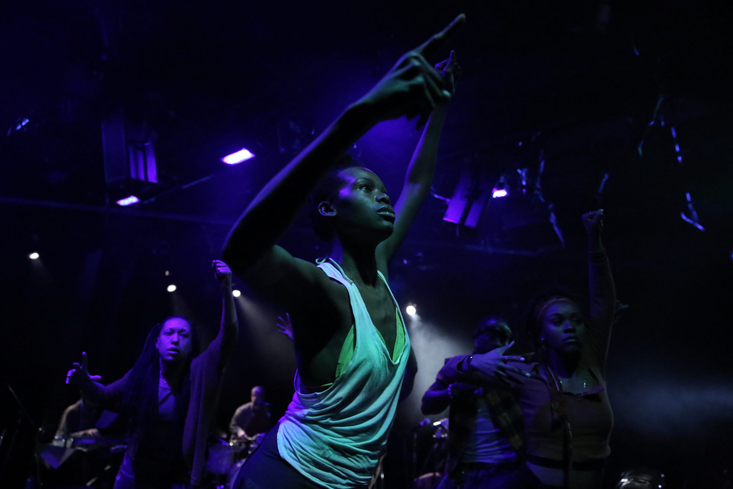 Chanon Judson is seen in a blacklight on a dark stage wearing a white tank top, looking up with her arms over her head.