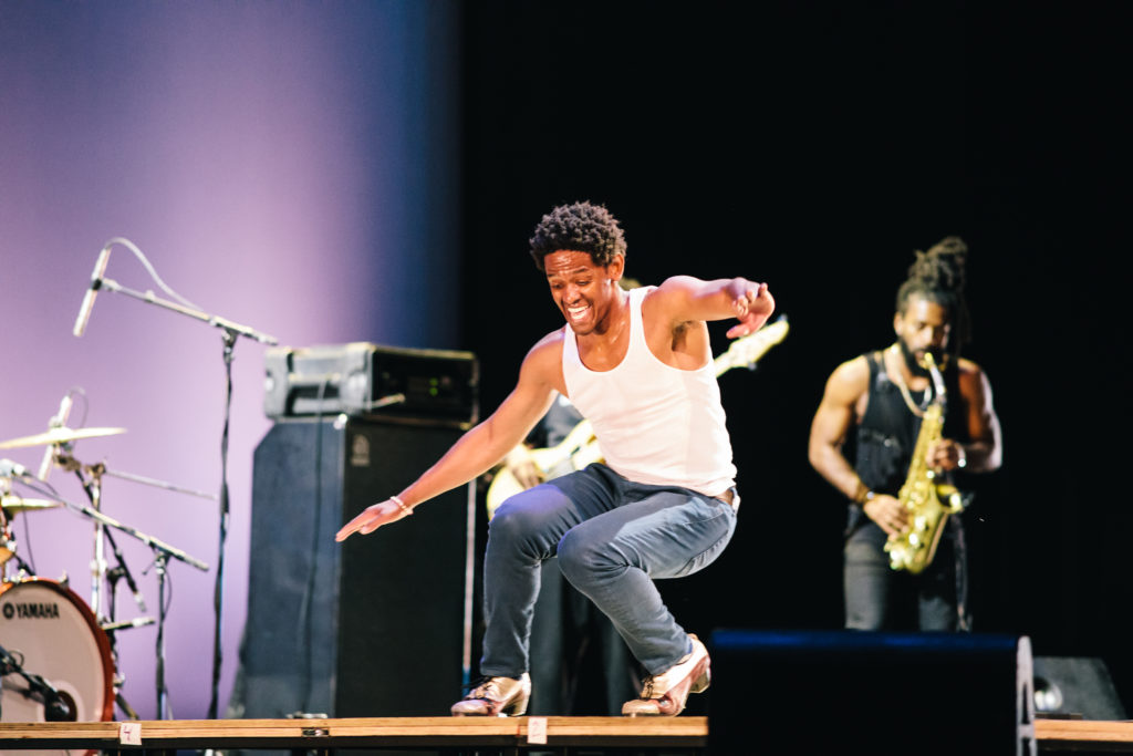 Jared Grimes grins as he balances in a deep crouch, arms outstretched for balance. He's on a wooden tap board onstage, wearing tap shoes and a white tank top, brow glistening with sweat. Musicians wielding a sax and bass guitar are blurry behind him.