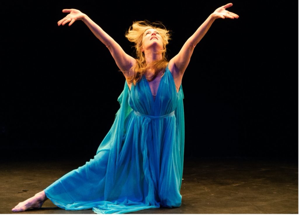 A woman in a diaphanous blue dress kneels with one leg extended side. Her arms form a V overhead, palms upturned, as she gazes up into a stage light, blonde hair flying.