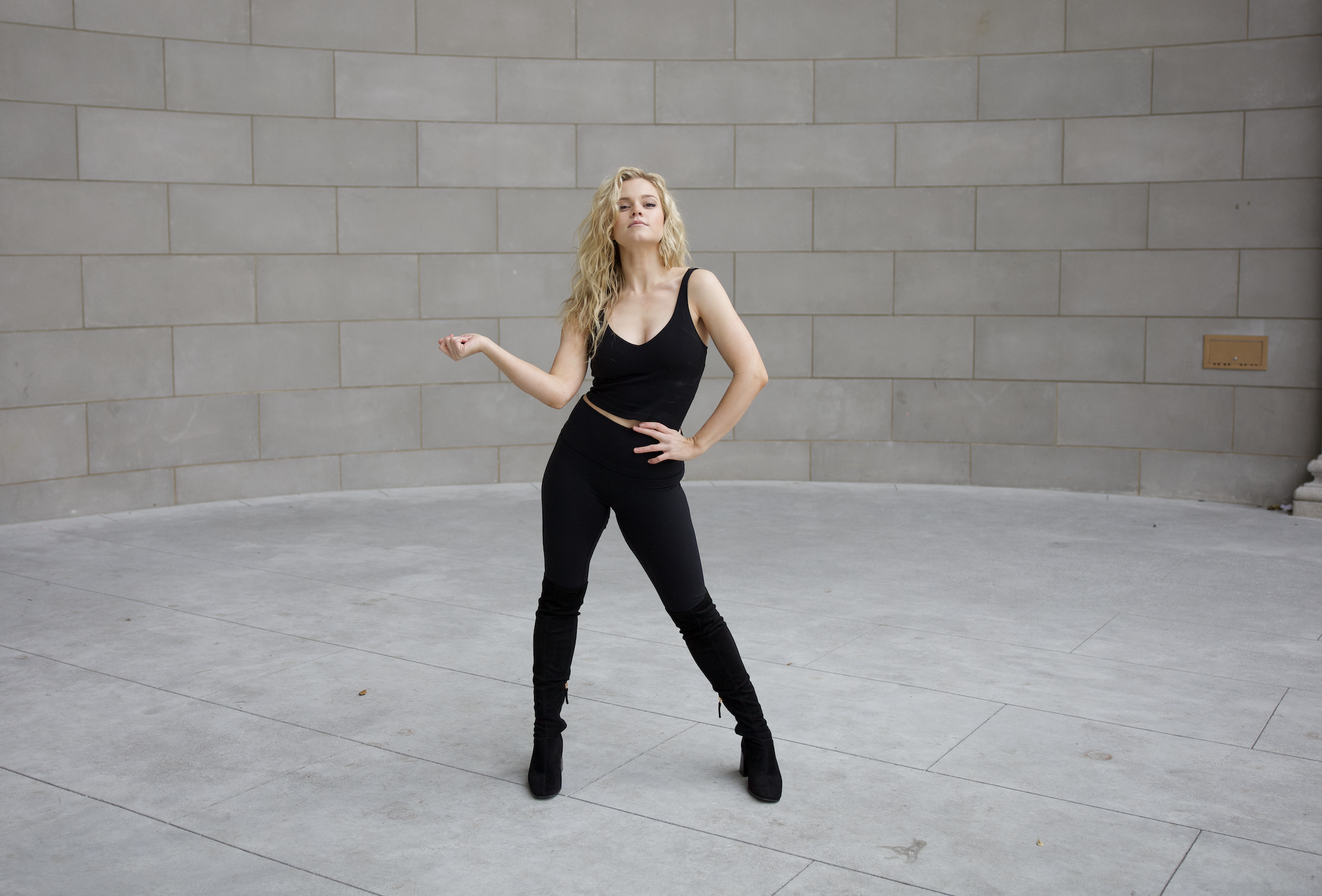 A blond woman stands with one hand on her hip, the other snapping to the side, in balck against a stone background