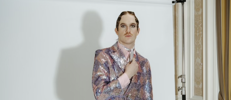 Austin Goodwin, a white man in a colorful suit, stands in front of a makeshift white backdrop, holding his lapel and looking resolutely into the camera