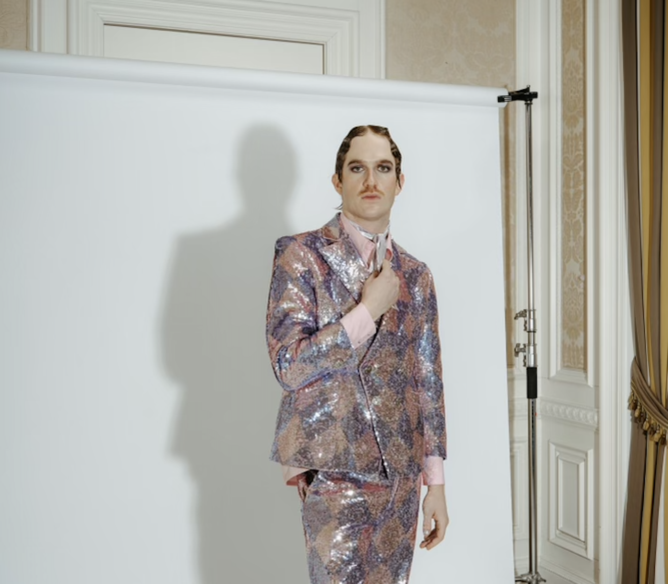 Austin Goodwin, a white man in a colorful suit, stands in front of a makeshift white backdrop, holding his lapel and looking resolutely into the camera