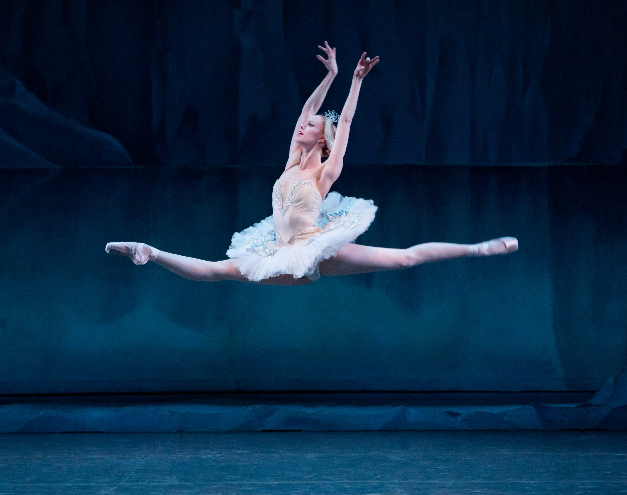 Teresa Reichlen, in costume as Odette, is captured mid-leap. Her legs are at a precise 180 degrees, parallel to the stage, and her arms curve up on either side of her ears, wrists almost touching overhead.