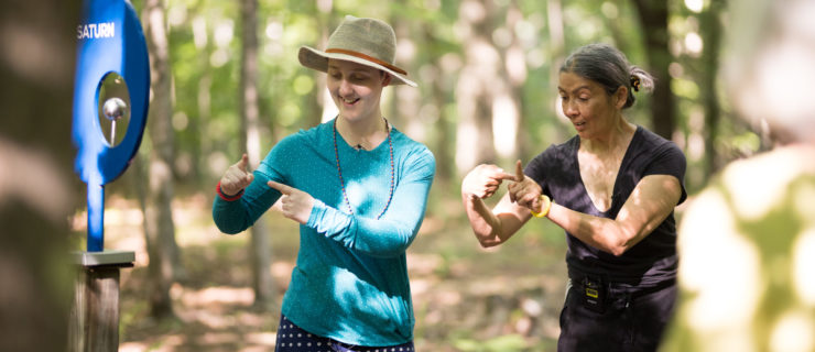 Standing in the woods during a Big Move at the Hop event, Emmanuèle Phuon speaks as she looks to where she is bringing her pointer fingers together. A woman in bright street clothes and a hat smiles as she imitates the gesture beside her.