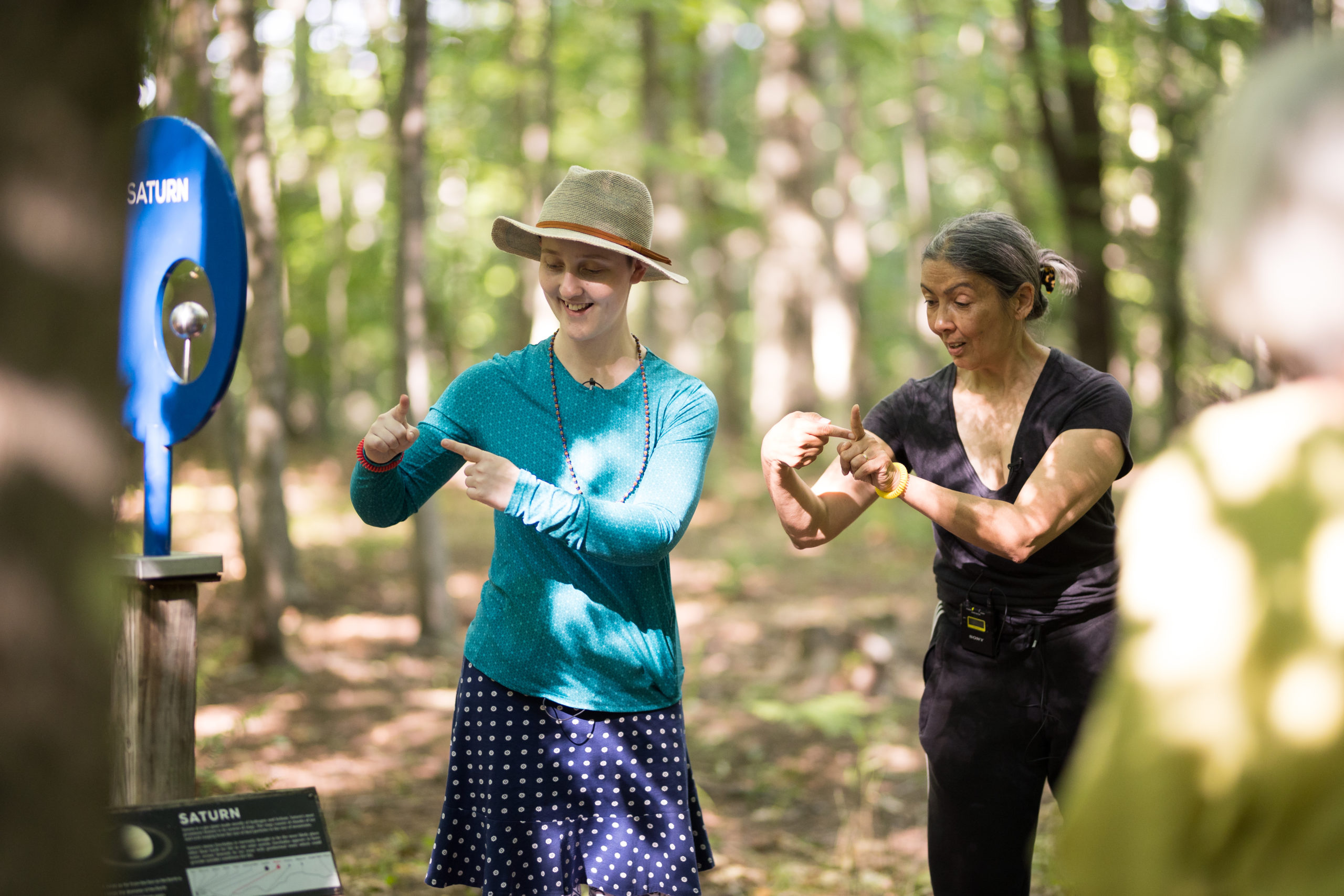 Standing in the woods during a Big Move at the Hop event, Emmanuèle Phuon speaks as she looks to where she is bringing her pointer fingers together. A woman in bright street clothes and a hat smiles as she imitates the gesture beside her.