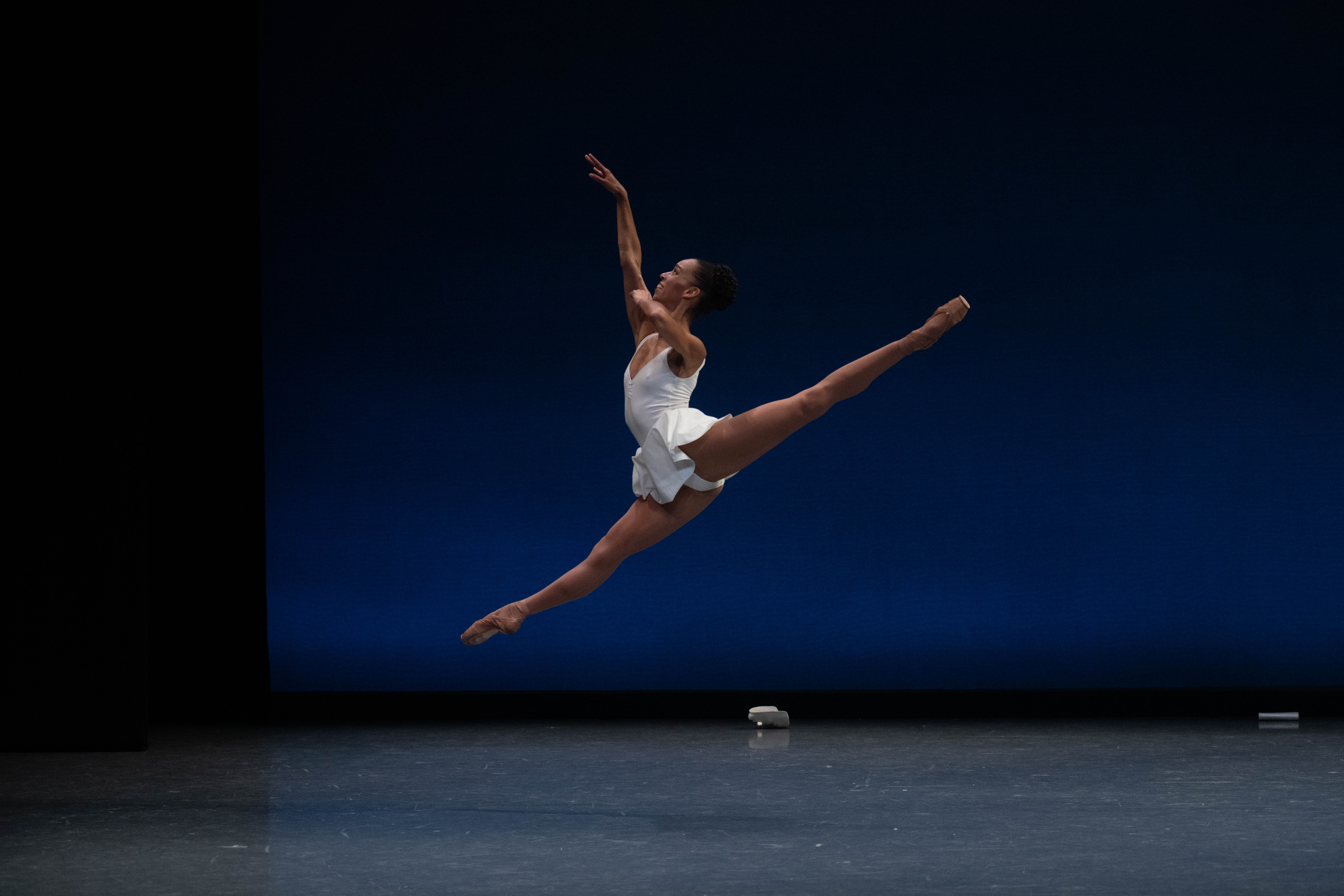 On a bare stage, Chyrystn Fentroy is caught mid-leap, her back leg above 90 and her front leg just below. Her downstage hand touches her upstage shoulder, with the upstage arm extended high. She wears a white tunic over tights and pointe shoes matching her skin tone.