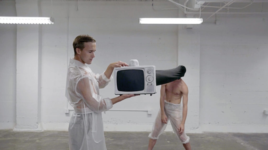 One man holds a fake TV with a long pipe covering another man's head. They are in an empty white room.