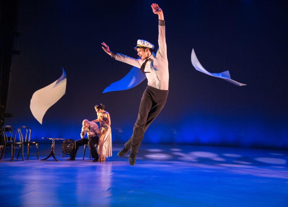 Dancer Zachary Kapeluck soars through the air onstage in a leap, both legs straight below him in a fourth position. Fabric swirls through the air around him.