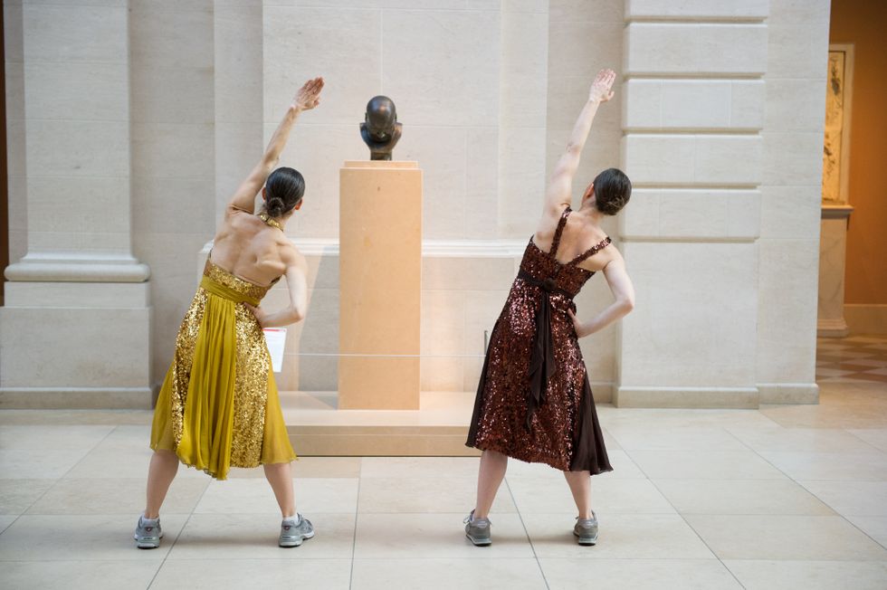Two women in sparkly dresses and sneakers stretch to their right side with their left arm up in the air in front of a statue.