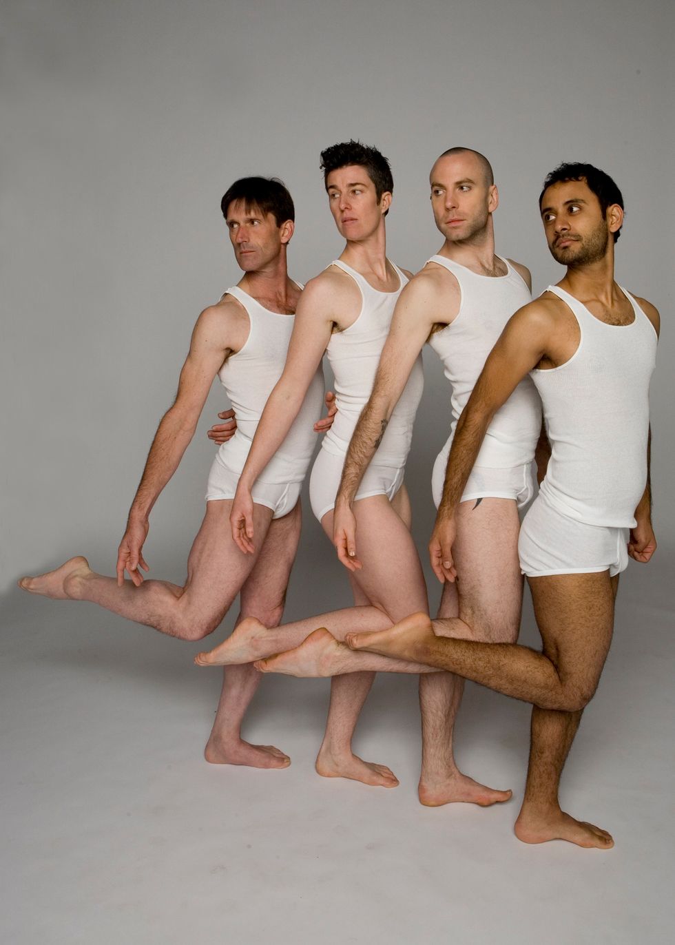 Four dancers in white tank tops and briefs stand close together with their working legs bent, looking back.