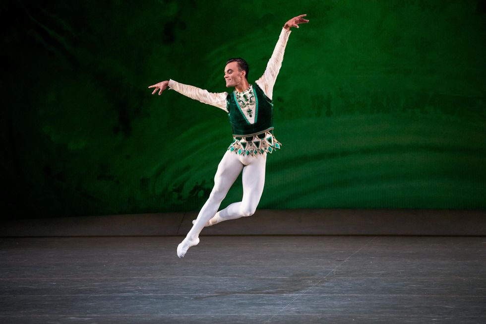 Stanley flies through the air in jete side, a green background at the back of the stage matching his green vest over white tights.