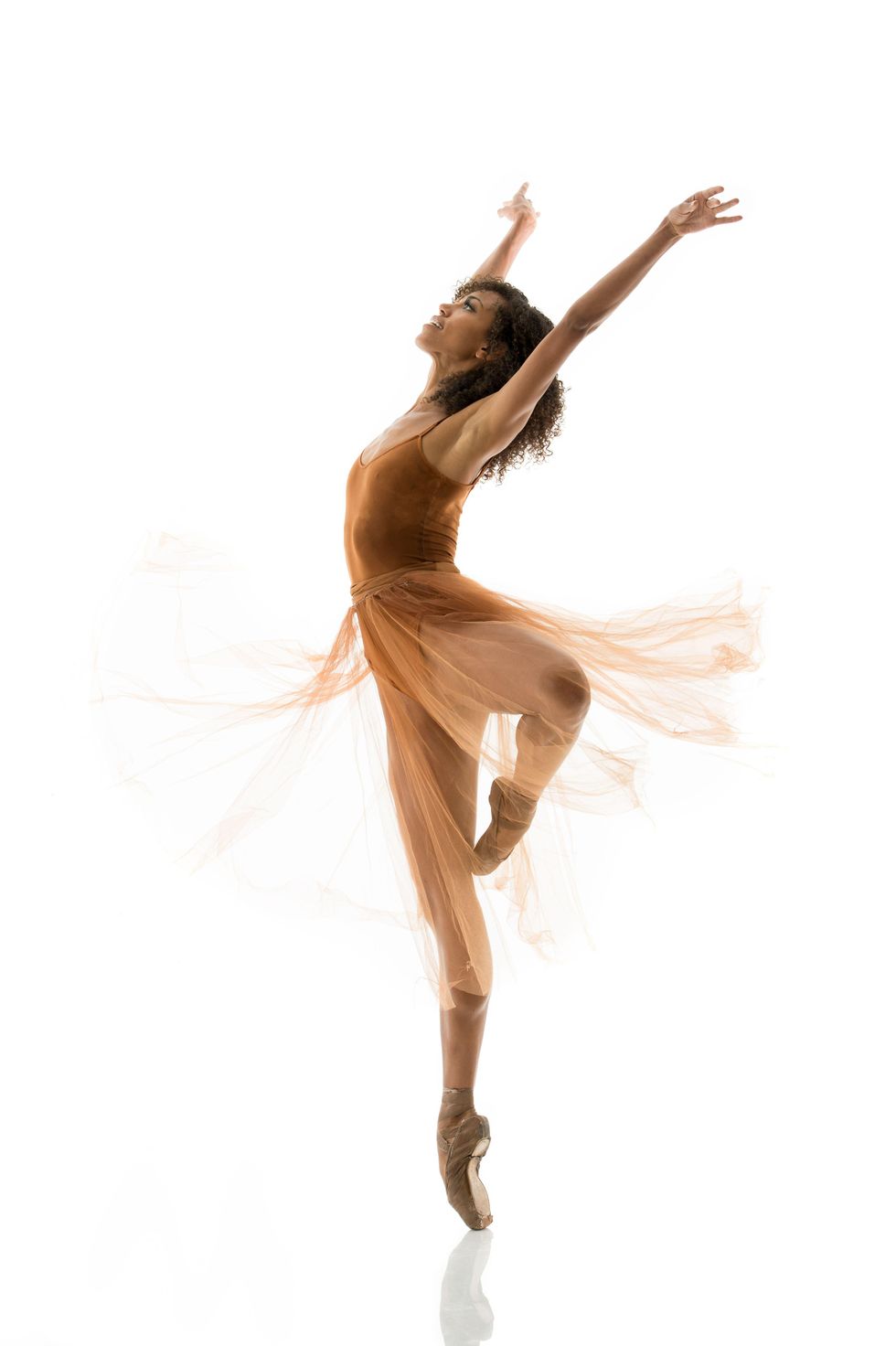 Donnell stands on pointe in passe back, her hands reaching up and behind her head, in an orange dress that flows behind her.