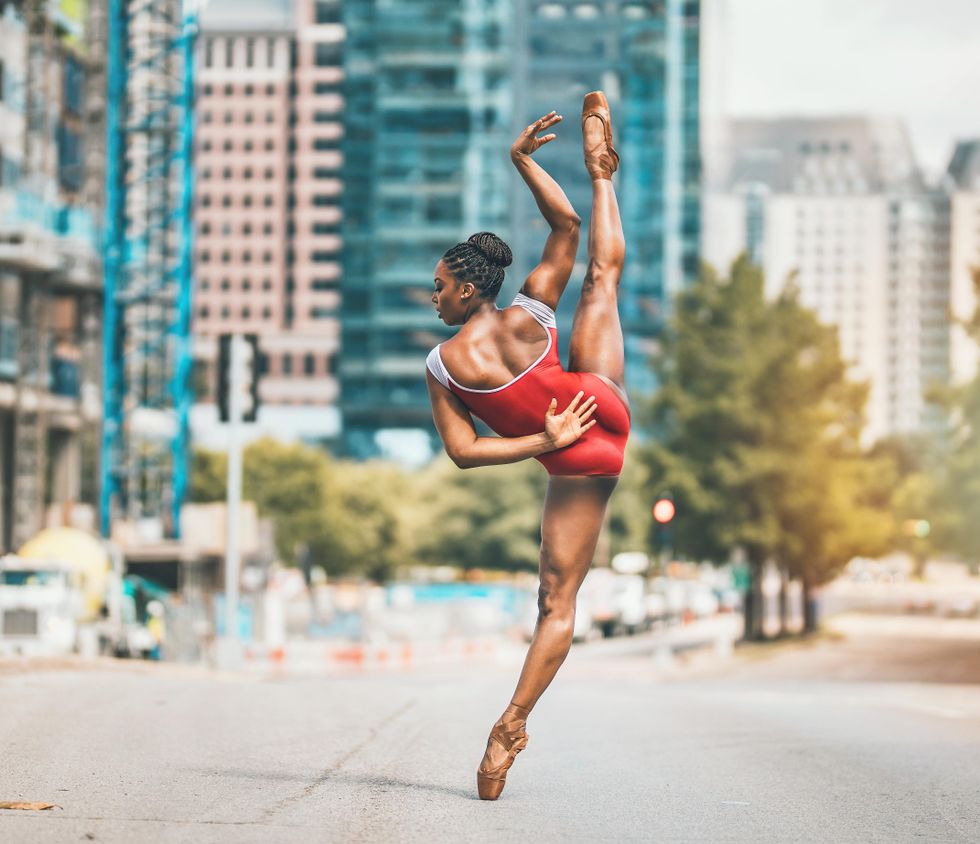 Newkirk stands on pointe in just a leotard and brown pointe shoes, one leg developpe side, facing with her back to us on an empty city street