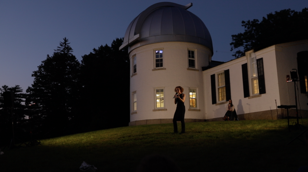 As night falls, Emily Coates speaks into a microphone on the grass in front of a round, white observatory building, which houses
