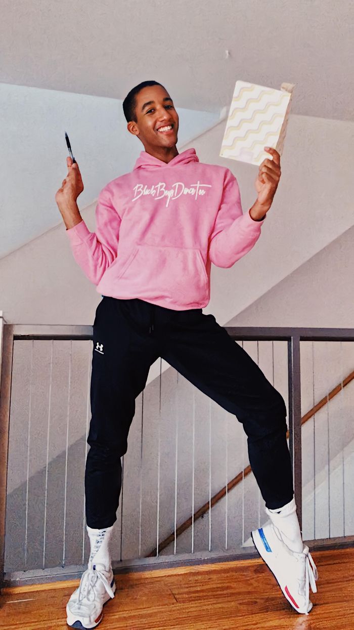 Watters, a Black man with short hair, stands in front of a staircase. He wears white sneakers, black sweatpants and a pink sweatshirt. He holds up a white journal and a pen.