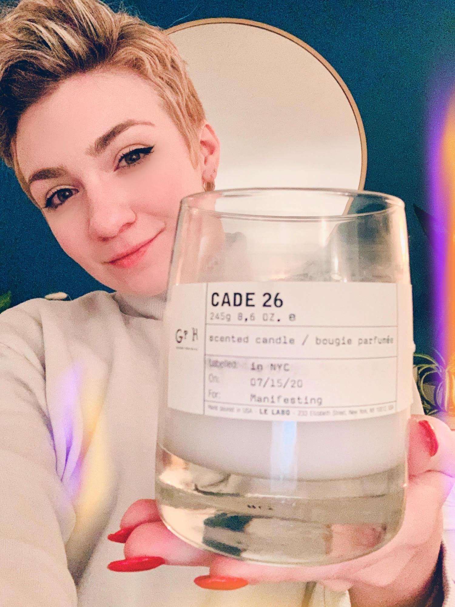Garcia-Lee, a white woman with short blonde hair, holds a candle labeled "Cade 26" in her hand. She smiles, has bright red nail polish on, and wears a light-colored sweatshirt.