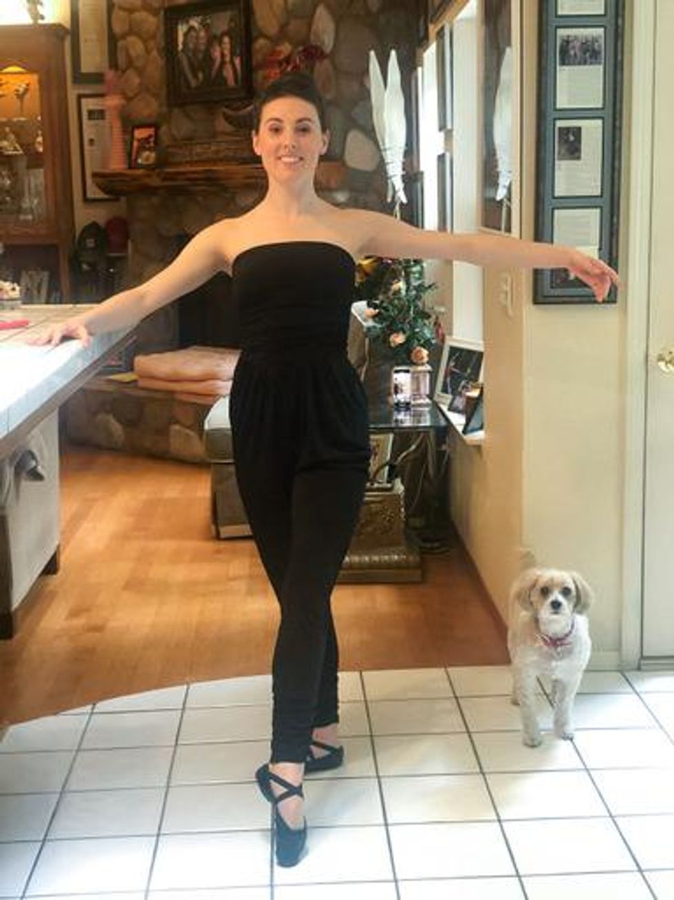 Peck, a woman with brown hair, stands in a tendu ballet position with one foot pointed in front of her on the floor. One hand holds her countertop for support and the other is to the side of her in second position. She wears a black jumpsuit and black ballet slippers. She stands on white tile next to a white dog. A brick fireplace is behind her.