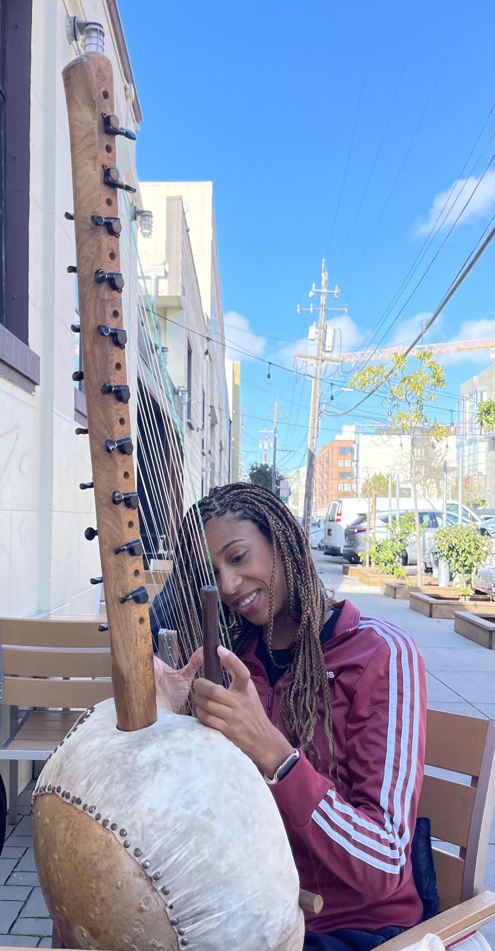 Cissoko, a Black woman with light brown braids, sits in a chair and plays the kora, a lute-like string instrument made of wood and leather. She wears a red jacket and sits outside on a sidewalk.